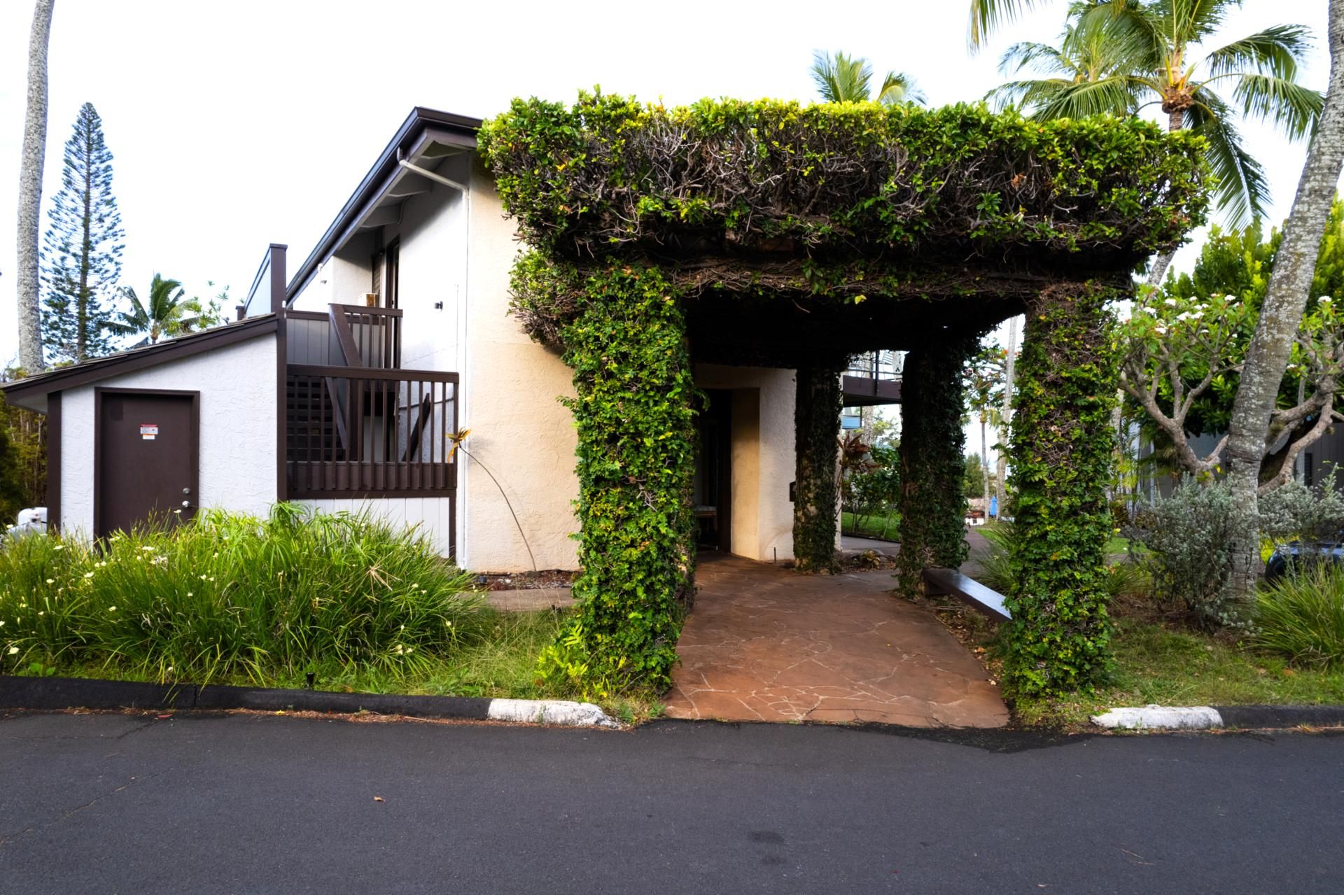 Ein Haus mit überdachter Veranda, einer kleinen Terrasse und einem Garten mit unterschiedlichen Pflanzen und einer Palme.