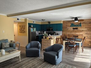 Une cuisine moderne avec des meubles de couleur turquoise, un coin repas avec des chaises en bois et un salon avec un canapé et des fauteuils.