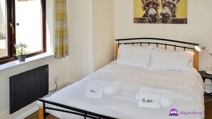 A neatly made bed with white linens and towels, a black metal headboard, a window with a curtain, and a wall-mounted painting of donkeys. - Station 1 Bed Apartment – Stayseekers (Wiltshire)