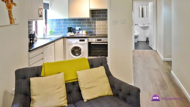 A compact living space with a grey sofa, yellow cushions, a kitchen with a washing machine, and a bathroom with a white sink. - Station 1 Bed Apartment – Stayseekers (Wiltshire)