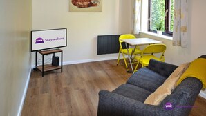 A furnished room with a sofa, a small table, a yellow chair, a TV on a stand, and a window with curtains. - Station 1 Bed Apartment – Stayseekers (Wiltshire)