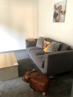 A grey sofa with two cushions, a small wooden table, and a brown animal-shaped ottoman in a room with a plain white wall and a framed picture.