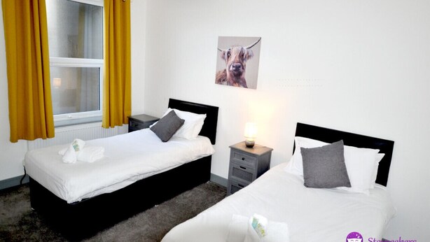 Hotellihuone, jossa on kaksi sänkyä, ikkuna, jossa on keltaiset verhot, yöpöytä lampun kanssa ja seinälle ripustettu kuva koirasta.