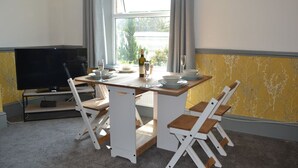 Un coin repas avec une table dressée pour quatre personnes, un téléviseur posé sur un meuble et une fenêtre avec des rideaux.
