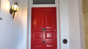 Uma porta vermelha com moldura branca, uma pequena janela acima dela e uma luminária fixada na parede à esquerda.