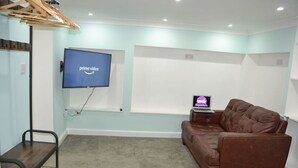 A room with a large flat-screen TV mounted on the wall, a brown leather sofa, a small table with a laptop, and a wooden shelf.