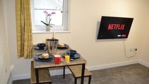 A dining area with a table set for two, a TV mounted on the wall displaying the Netflix logo, and a window with curtains and a potted plant. - Olive Tree 2 bed Apartment - Stayseekers (Salisbury)