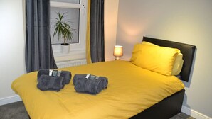 A neatly made bed with a yellow bedspread, two rolled towels, and a bedside lamp. - Olive Tree 2 bed Apartment - Stayseekers (Salisbury)