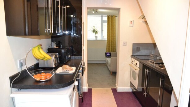 A compact kitchen with a black countertop, a sink, and a stove. There is a window with a curtain, a plant, and a view into a living area. - Olive Tree 2 bed Apartment - Stayseekers (Salisbury)