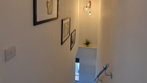 En smal trappa med en lampa högst upp, en liten växt på en hylla och inramade tavlor på väggarna.