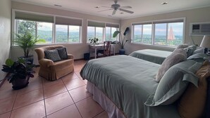Un dormitorio con cama, dos mesitas de noche, una silla y un escritorio. Amplias ventanas ofrecen vistas al exterior.