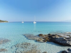 Mar turquesa y transparente con dos veleros anclados a lo lejos, costa rocosa en primer plano y cielo despejado.