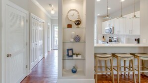 Un couloir avec des portes blanches, une étagère intégrée ornée d’objets décoratifs et un coin cuisine avec des meubles blancs et des tabourets de bar.