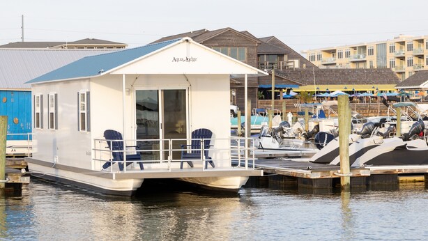 Una casa sull’acqua galleggiante con terrazza coperta e area salotto, ormeggiata in una marina con altre imbarcazioni e edifici sullo sfondo.