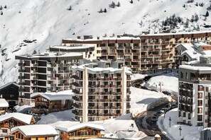 A snow-covered mountain resort with multi-story buildings, balconies, and a ski slope in the background.