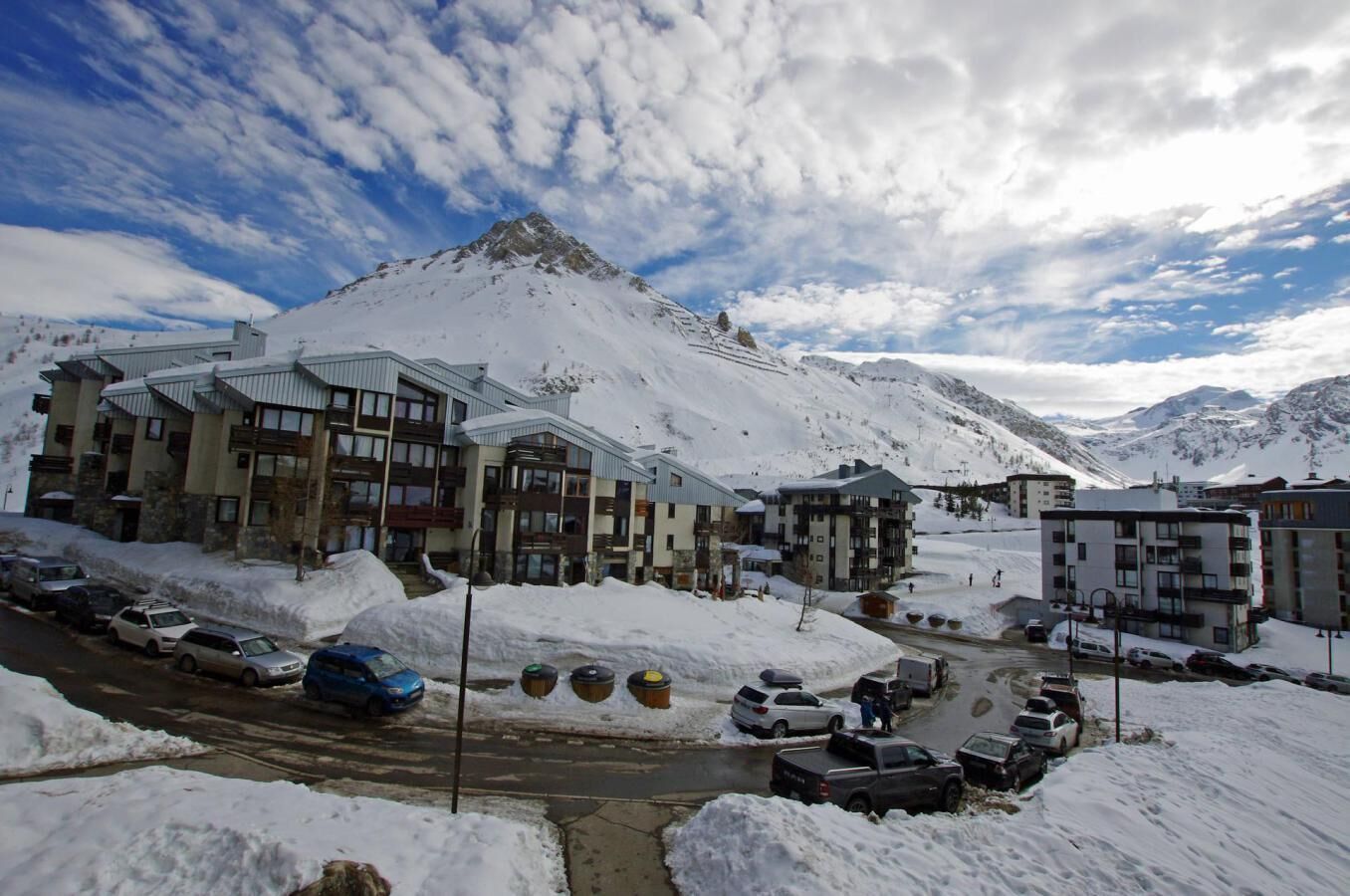 Un pueblo nevado en la montaña con edificios modernos, una carretera y coches aparcados.