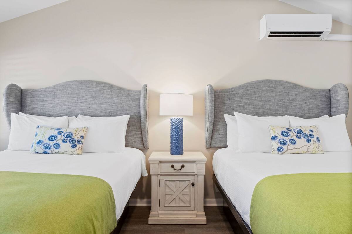 Habitación de hotel con dos camas, una mesita de noche con lámpara y un equipo de aire acondicionado.