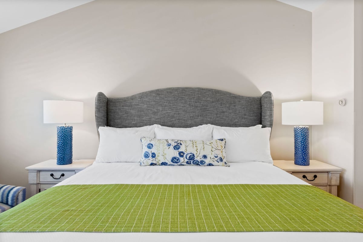 Una cama bien hecha con colcha verde a rayas, cabecera tapizada de gris y un cojín floral. Hay dos mesitas de noche, cada una con una lámpara y un jarrón azul.