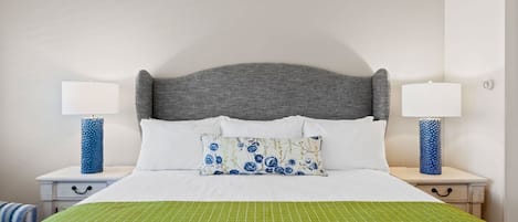 Una cama bien hecha con colcha verde a rayas, cabecera tapizada de gris y un cojín floral. Hay dos mesitas de noche, cada una con una lámpara y un jarrón azul.