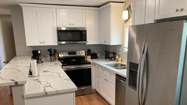 Una cocina compacta con gabinetes blancos, un microondas de acero inoxidable, horno integrado, un fregadero y un refrigerador.