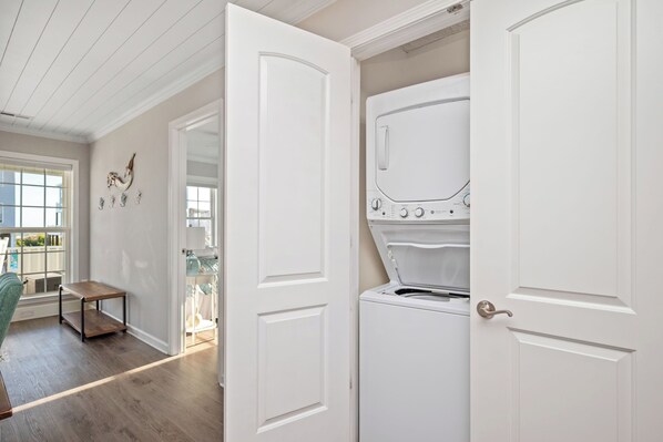 A laundry room with a white washing machine and dryer, a small wooden table, and a door leading to another room. - Mermaid Manor: Ocean View Home with Private Pool, Close to Beach Access (Oak Island)