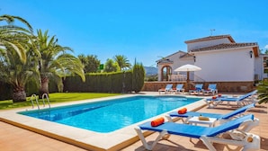 Une piscine avec des chaises longues et, au fond, un bâtiment blanc doté d’un toit recouvert de tuiles.