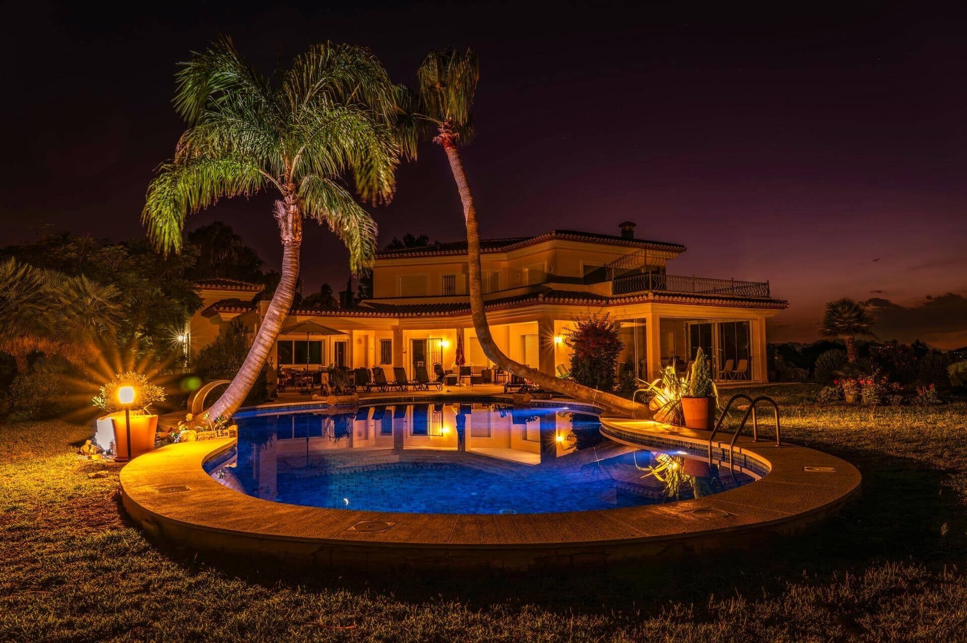 Une vue nocturne d’une maison luxueuse dotée d’une piscine, de palmiers et d’un espace salon extérieur.