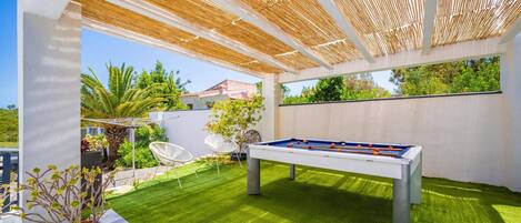 An outdoor pool table with a clear view of the surrounding greenery and a white wall.