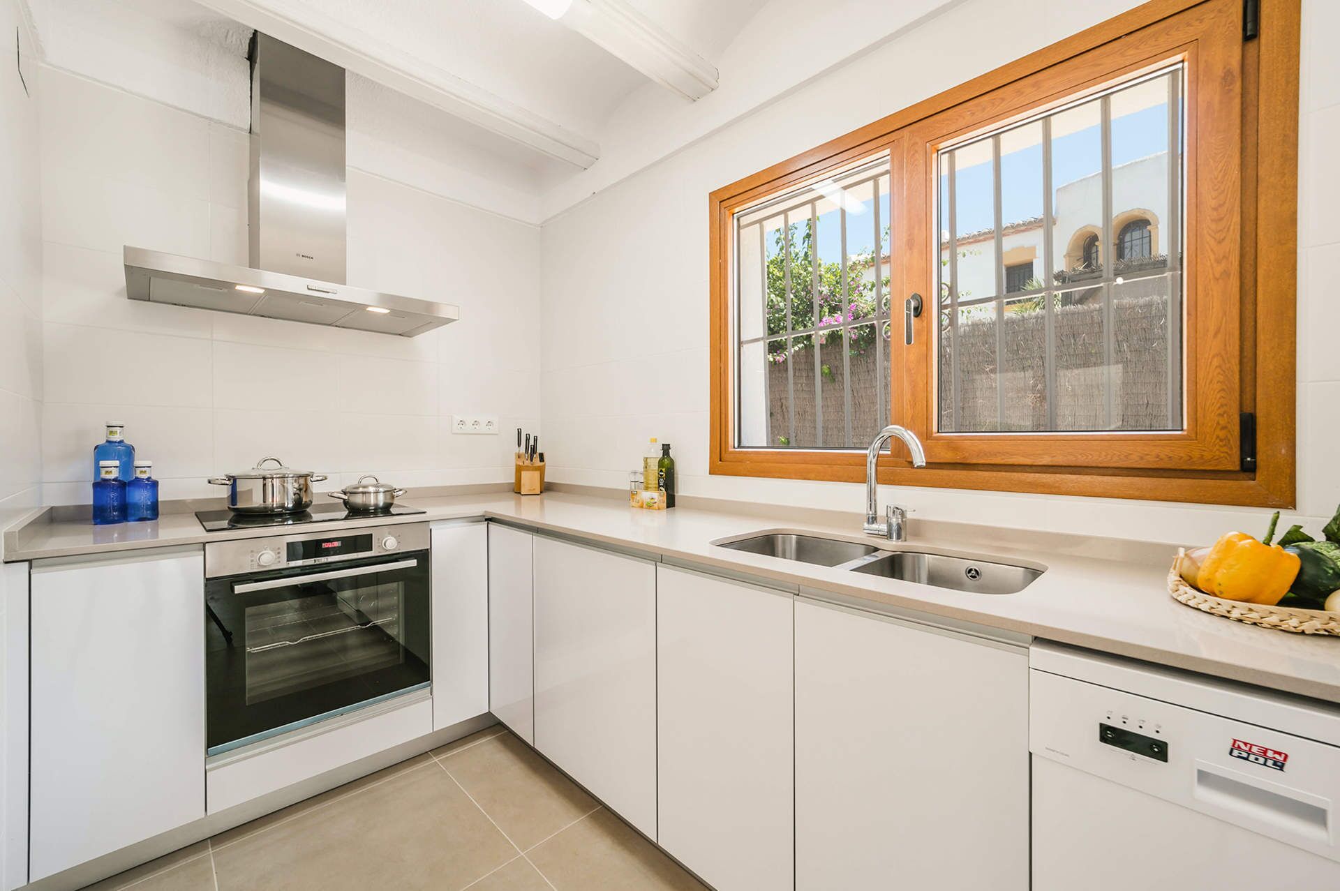 Una cocina moderna con armarios blancos, una estufa de acero inoxidable, un horno, un lavavajillas y un ventanal con vistas a un jardín.