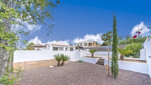 Une maison moderne à l’extérieur blanc, avec une allée en gravier et un petit jardin agrémenté de palmiers et d’un grand conifère.
