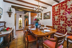 A dining area with a wooden table set for four, surrounded by chairs with red cushions. A chandelier hangs above, and a vase with flowers is on the table.