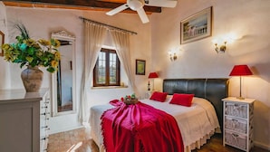 Une chambre à coucher comprenant un lit, deux lampes de chevet, une fenêtre avec des rideaux, un ventilateur de plafond et un tableau accroché au mur.