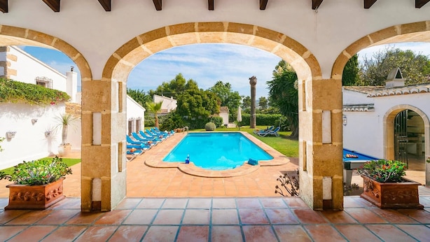 Une piscine à l’eau bleue, entourée d’une terrasse carrelée et située sous une zone abritée avec des arcades.