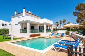 Una casa moderna de dos pisos amb una piscina, espais d'exterior per seure i un cel blau clar.