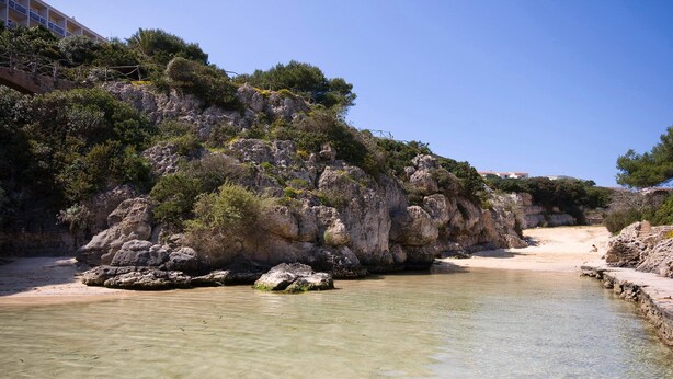 A coastal area with clear shallow waters, rocky cliffs, and a sandy beach.