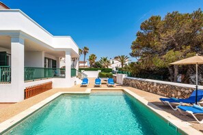 Zona de piscina con una piscina azul cristalina, sillas reclinables azules y un edificio blanco con balcón.