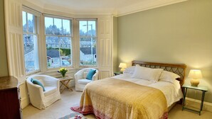Un dormitorio con una cama grande, dos sillas blancas, una mesita con flores y vistas al exterior a través de amplias ventanas.