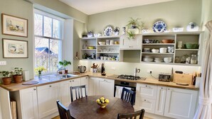 Una cocina con armarios blancos, encimera de madera y una mesa de comedor con sillas. Hay plantas en macetas sobre la encimera y estantes con vajilla y frascos.