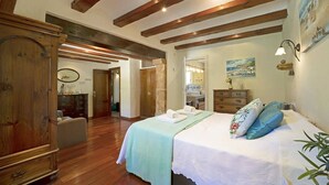 Une chambre avec des poutres en bois, un lit agrémenté d’un coussin à motifs floraux, une commode et un tableau représentant une scène de plage.