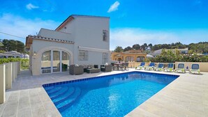 A swimming pool with a tiled edge, a covered seating area with chairs, and a building with an arched entrance. - Villa Toscamar by Villa Plus (El Tosalet)