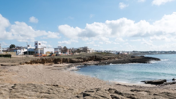 Una zona costera con playa de arena, costa rocosa y edificios al fondo.