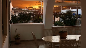 Un balcón con vistas a un atardecer, una mesa redonda con sillas y plantas en macetas.