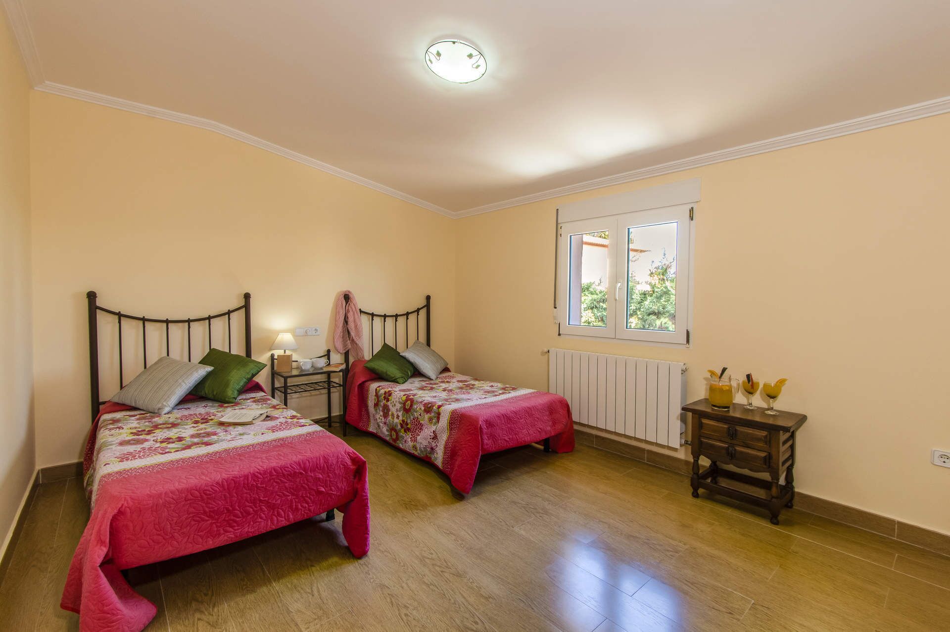 A bedroom with two beds, a bedside table with a lamp, a window with a view of greenery, and a small wooden side table with drinks.