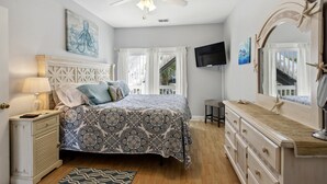 Quarto com cama, penteadeira, espelho, ventoinha no teto e televisão na parede.
