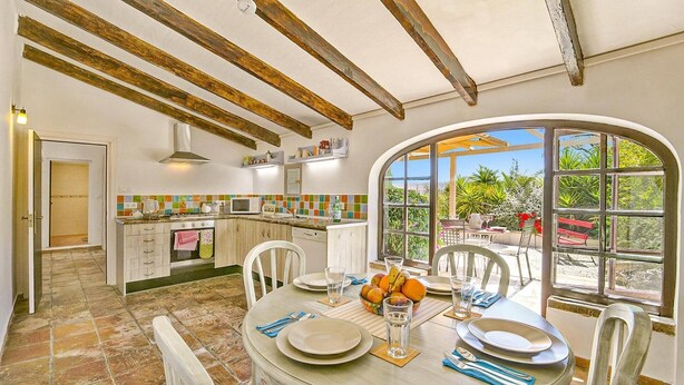 A dining area with a round table set for four, a kitchen in the background, and a view of greenery through a large window. - Villa Jessie by Villa Plus (Montgo de Toscamar)