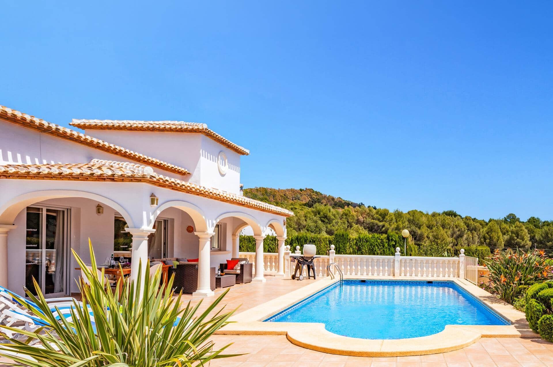 Área de piscina com um edifício branco, assentos externos e vista clara para uma encosta.