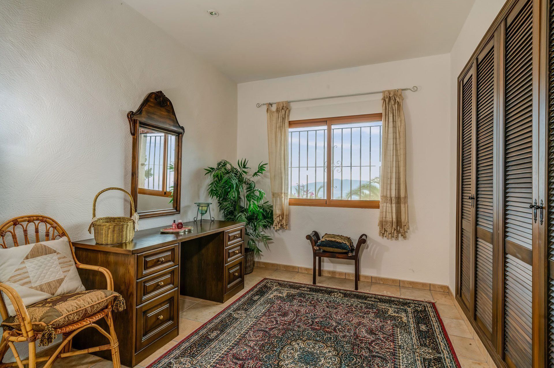 A room with a desk, wicker chair, and a large mirror. There is a window with curtains and a view of the outdoors.