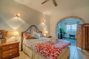 A bedroom with a bed, bedside lamps, a wooden dresser, and a view through an arched doorway to an outdoor area.