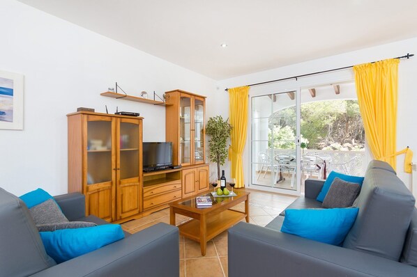 A living room with a sofa set, a wooden cabinet with a TV, a small table with a plant and wine glasses, and a sliding glass door leading to an outdoor area.