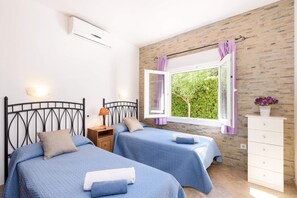 A bedroom with two beds, a window with purple curtains, a bedside table with a lamp, and a dresser.